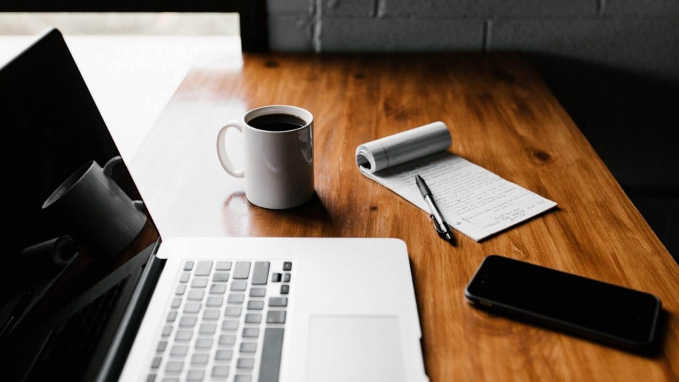 computer, mug, notepad, pen, and phone on a tabletop