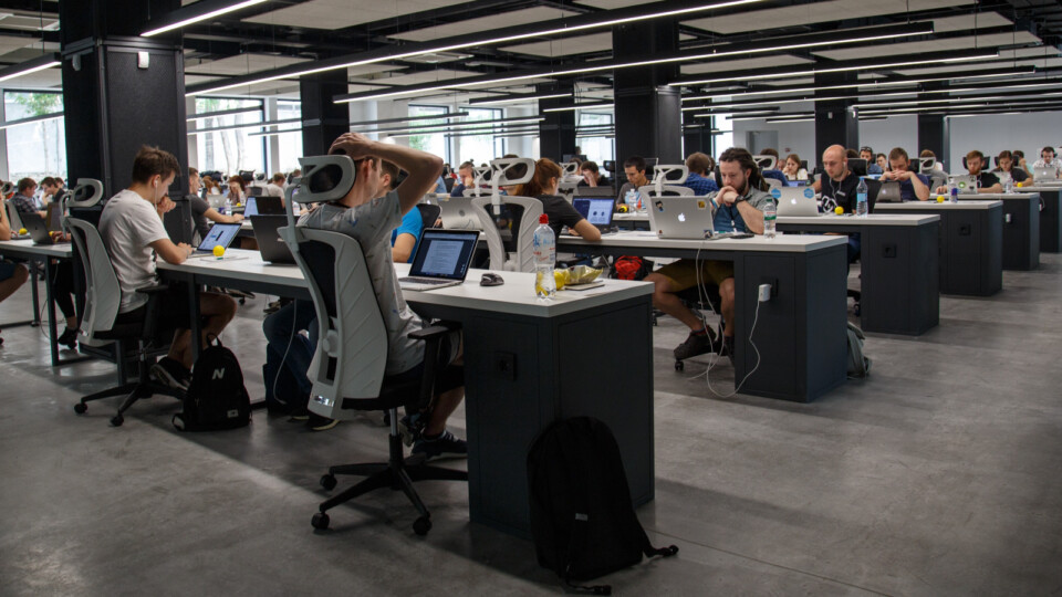 Um escritório com diversas mesas longas cheias de pessoas trabalhando lado a lado