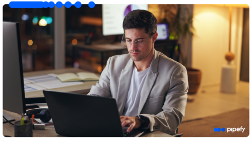 Homem jovem de óculos e blazer cinza trabalhando concentrado em um laptop em um escritório com iluminação quente ao fundo durante a noite, simbolizando a agilidade do RPA na execução de tarefas digitais