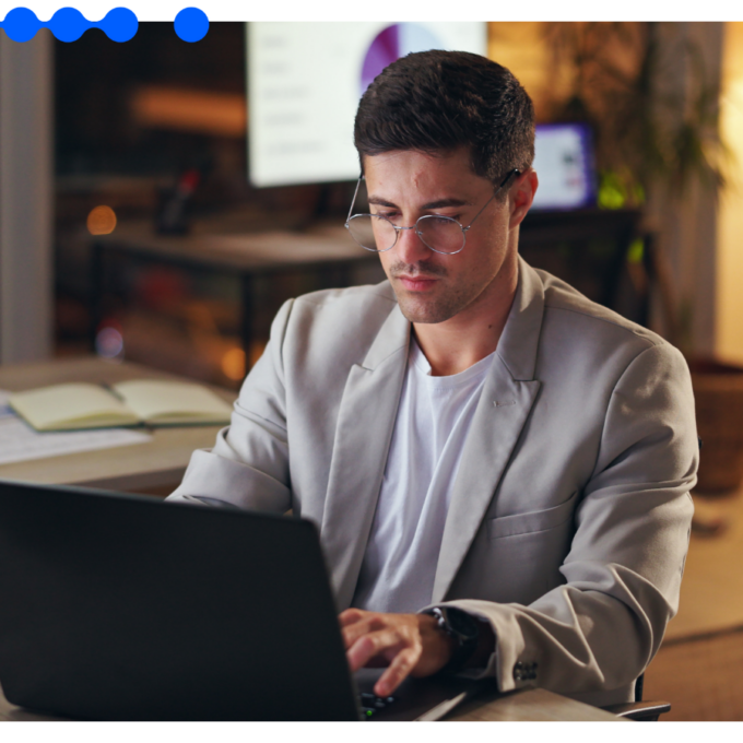 Homem jovem de óculos e blazer cinza trabalhando concentrado em um laptop em um escritório com iluminação quente ao fundo durante a noite, simbolizando a agilidade do RPA na execução de tarefas digitais