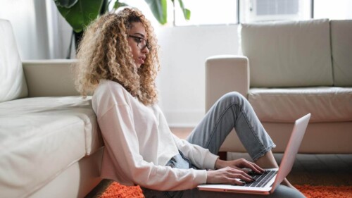 Moça de cabelo encaracolado sentada no chão em frente a um sofá usando um notebook.