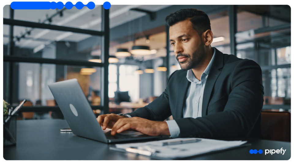 Homem executivo vestindo terno e camisa social trabalhando focado em um laptop em um escritório moderno, com papéis sobre a mesa, organizando o processo de vendas
