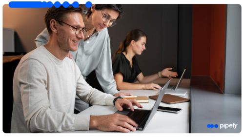 Two professionals in an office smiling and looking at a laptop screen together, discussing how to create a workflow, with another woman working in the background