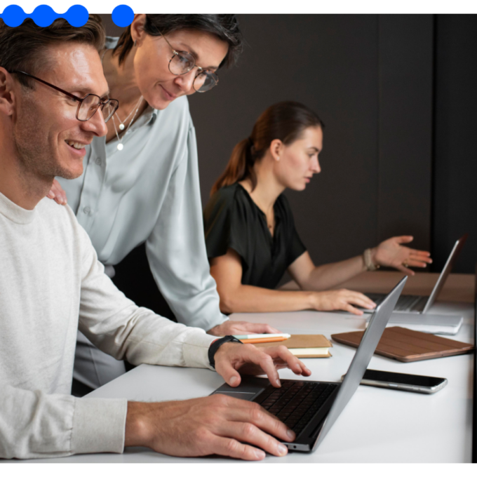 Two professionals in an office smiling and looking at a laptop screen together, discussing how to create a workflow, with another woman working in the background