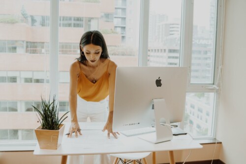 Mulher em pé organizando papéis próxima a um computador