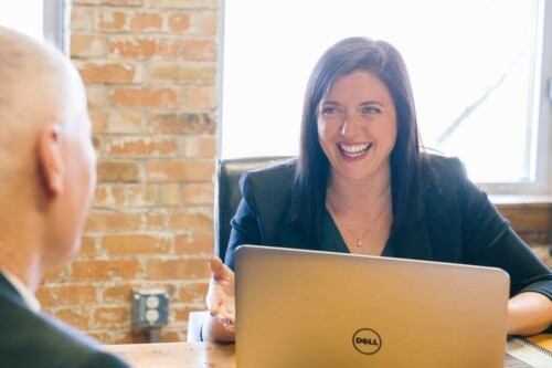 Uma moça sorridente usando terninho em frente a um notebook da Dell sentada em uma mesa num escritório com paredes de tijolos expostos.