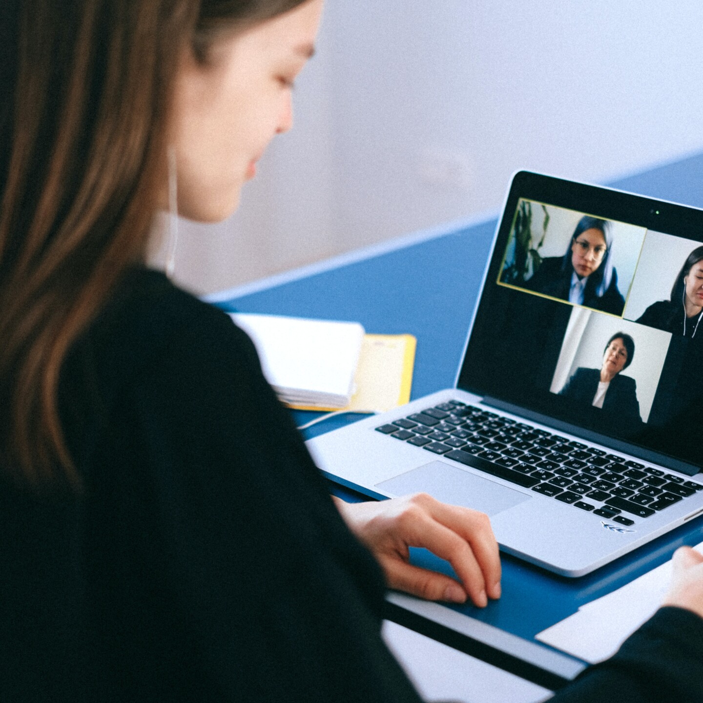 Moça realizando reunião remota pelo MacBook. Na tela dela vemos outras três moças, cada uma em uma janela diferente, todas trajando roupas formais. 