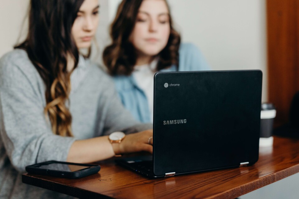 Duas moças em trajes de escritório trabalhando juntas em um notebook. Uma digita enquanto a outra, mais longe da tela, observa.