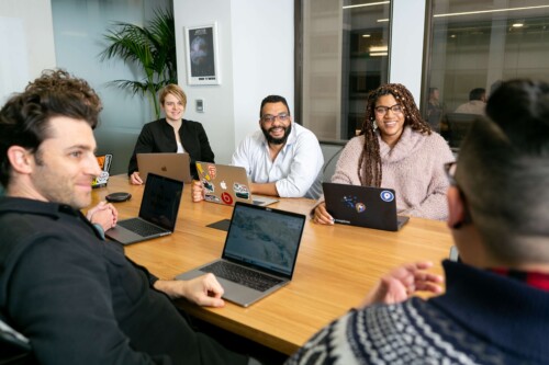 Grupo de colaboradores debate com laptops abertos em mesa de madeira