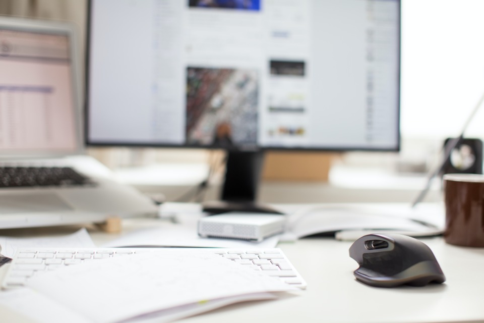 A desk with a keyboard, mouse, and notepad in focus. OUt of focus, further from the camera, there are also a laptop computer and a monitor.