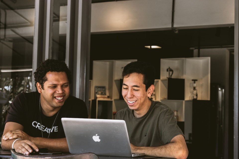 Two men in an office looking at a laptop computer and smiling at what's on the screen.