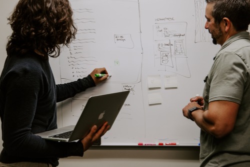 A female-appearing person stands with her back towards us, facing a whiteboard, writing on it with a marker with her left hand while holding her Apple notebook on her right arm. A male-appearing person stands to her right, looking at what she is writing.