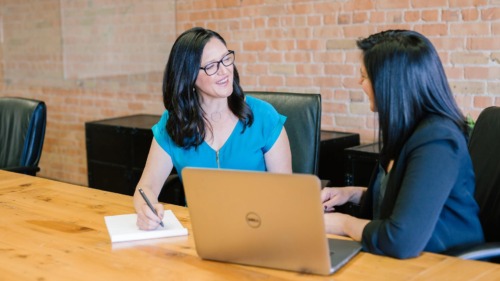 Two female-appearing people sitting side by side on a wooden table. The one on the left is writing something on a notepad with a pen while smiling at the one on the right, who was her left arm on the table in front of a notebook and smiles back.