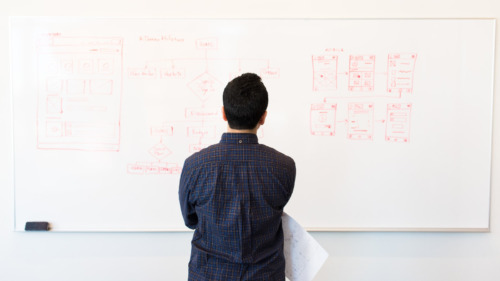 A male-appearing person with a dark blue shirt and short hair, his arms crossed in front of his chest, seen from behind as he stares at a whiteboard with workflow diagrams written on it.