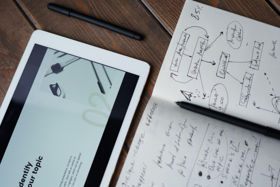 A tablet and a notebook on a wooden table. The tablet shows a website, while the notebook has a diagram written on it.