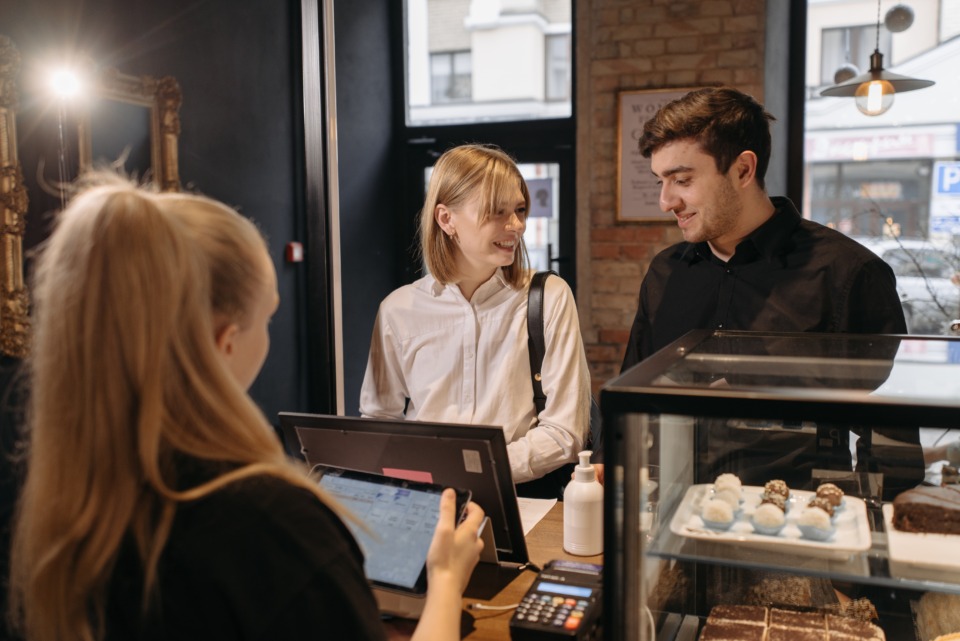 Uma moça loira e um homem branco no caixa de um café. A caixa, também branca, olha para os dois enquanto eles conversam entre si, pensando no que vão pedir