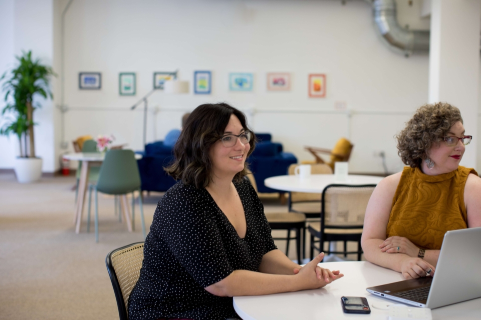 Duas mulheres brancas estão sentadas em uma mesa de trabalho com um laptop aberto