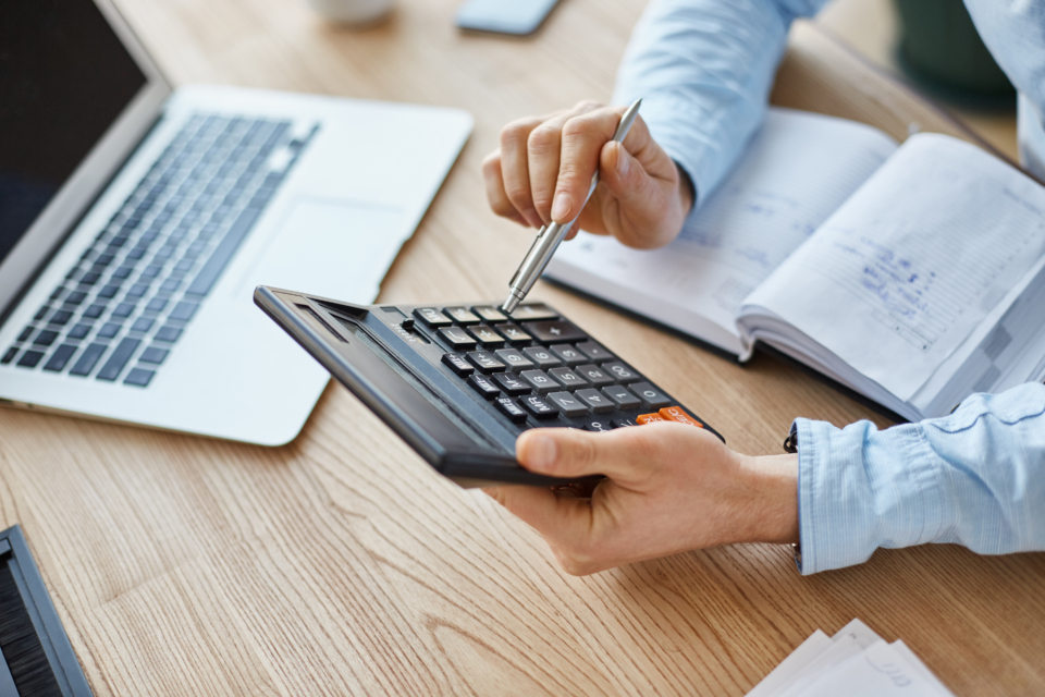 Homem com uma caneta na mão fazendo contas em uma calculadora. Atrás, um notebook aberto e um caderno com anotações.