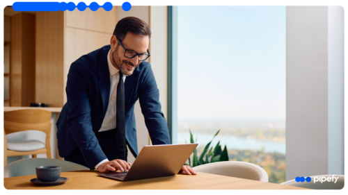 Homem sorridente de óculos e terno azul marinho inclinado sobre um laptop em uma mesa de madeira em um escritório bem iluminado com uma grande janela ao fundo, utilizando ferramentas no-code para otimizar seus processos de negócios