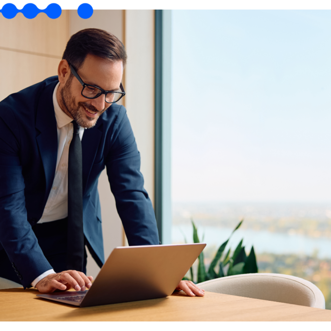 Homem sorridente de óculos e terno azul marinho inclinado sobre um laptop em uma mesa de madeira em um escritório bem iluminado com uma grande janela ao fundo, utilizando ferramentas no-code para otimizar seus processos de negócios