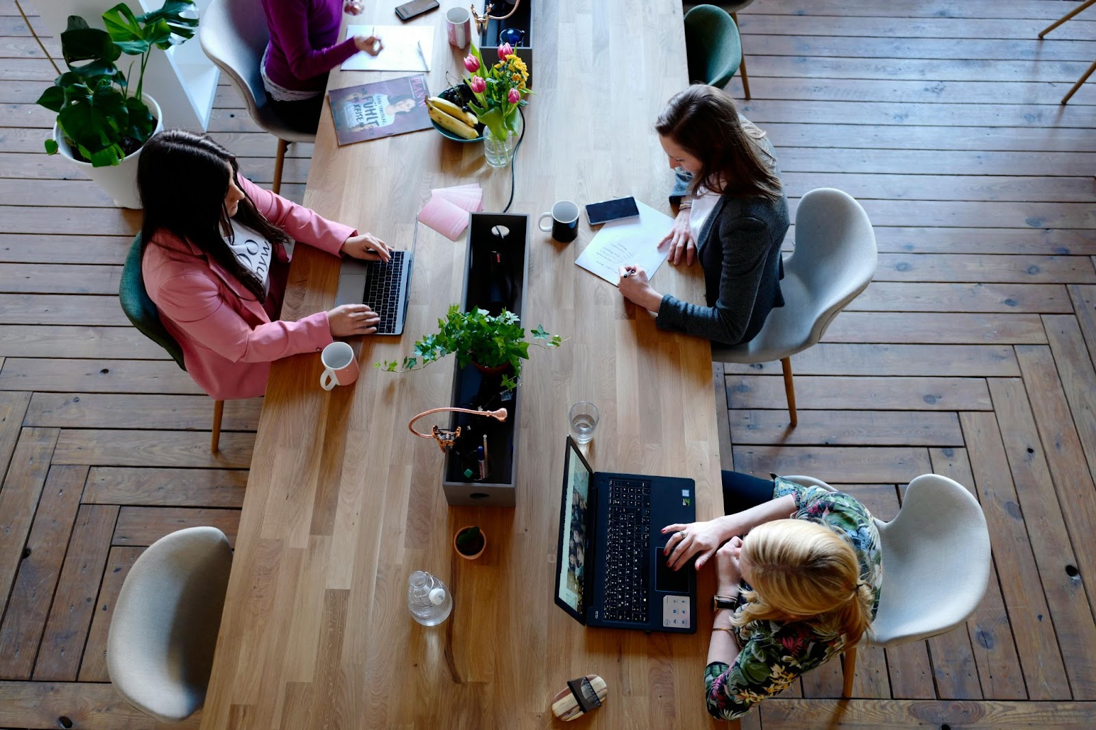 ao centro, uma mesa vista de cima. Ao redor da mesa, vemos 3 mulheres sentadas. Duas estão utilizando computador, enquanto a outra está escrevendo em uma folha.