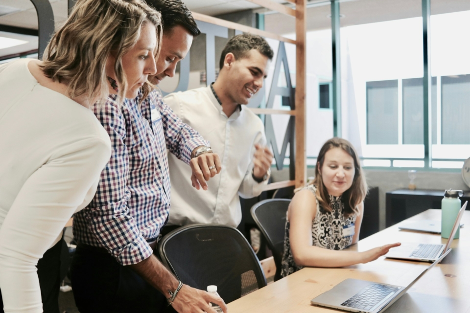 Four professionals collaborating at a table, focused on a laptop screen during a meeting in an office environment.