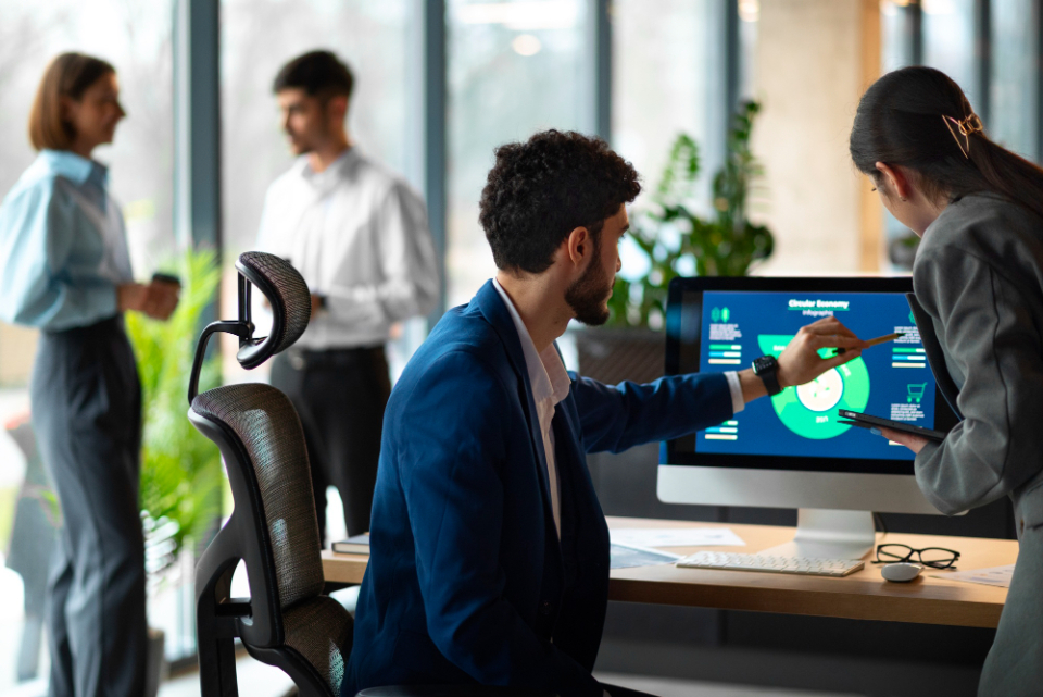 Two people collaborating in front of a computer displaying graphs on the screen while two others chat in the background, in a corporate setting.