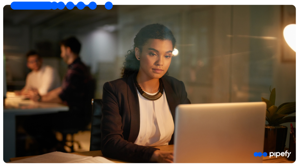 Mulher profissional focada trabalhando em um laptop em um escritório moderno à noite, realizando o processamento inteligente de documentos, com colegas reunidos ao fundo