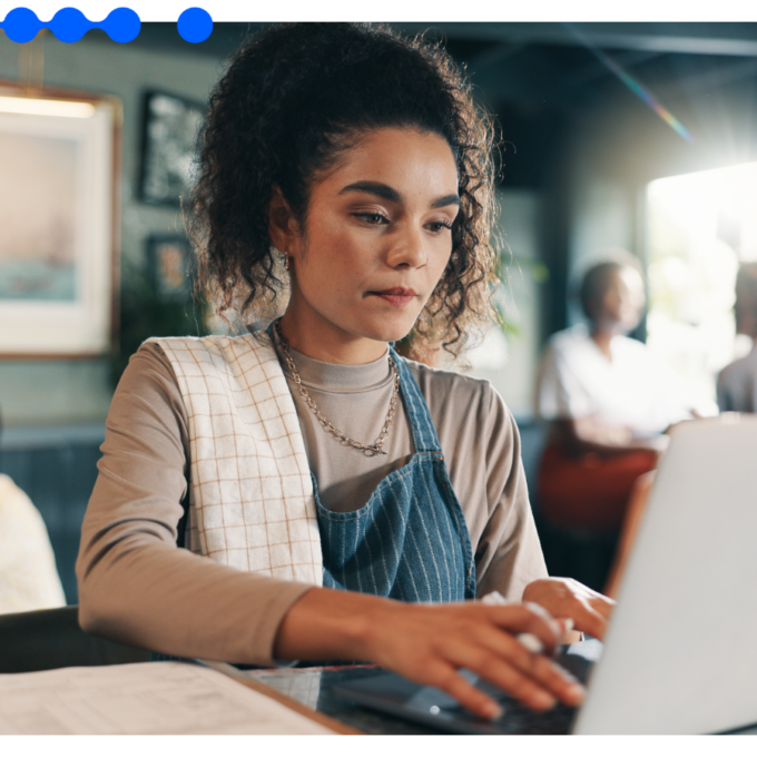 Mulher jovem trabalhando em um laptop em um café, usando avental e com uma toalha sobre o ombro, com outras pessoas ao fundo conversando
