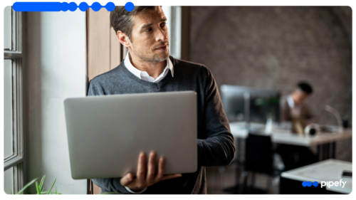 Homem adulto segurando um notebook enquanto olha pela janela em um escritório moderno, com outro profissional trabalhando ao fundo.