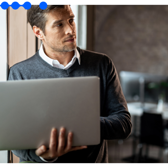 Homem adulto segurando um notebook enquanto olha pela janela em um escritório moderno, com outro profissional trabalhando ao fundo.