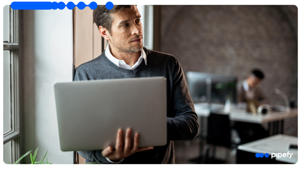 Homem adulto segurando um notebook enquanto olha pela janela em um escritório moderno, com outro profissional trabalhando ao fundo.