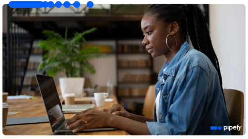 Mulher trabalhando em um laptop em ambiente de escritório, concentrada na tela, com estante de livros ao fundo e logotipo da Pipefy no canto inferior direito