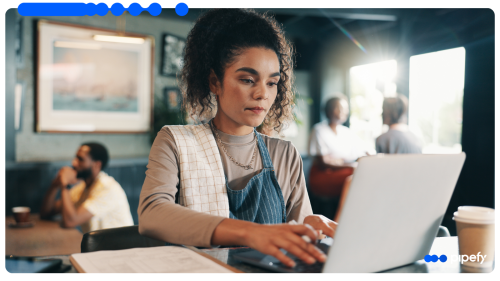 Mulher jovem trabalhando em um laptop em um café, usando avental e com uma toalha sobre o ombro, com outras pessoas ao fundo conversando.