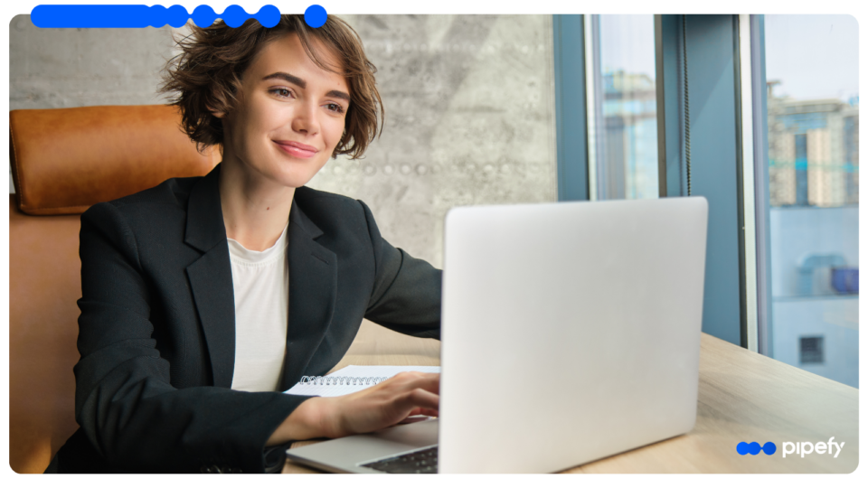 Professional woman in a dark blazer smiling while working on a laptop in a bright, modern office setting, representing a business leader researching what is process orchestration to improve organizational efficiency