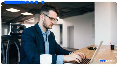 Homem jovem de óculos e blazer trabalhando em um laptop em um escritório moderno e iluminado, focado na análise de dados para prevenção de riscos operacionais