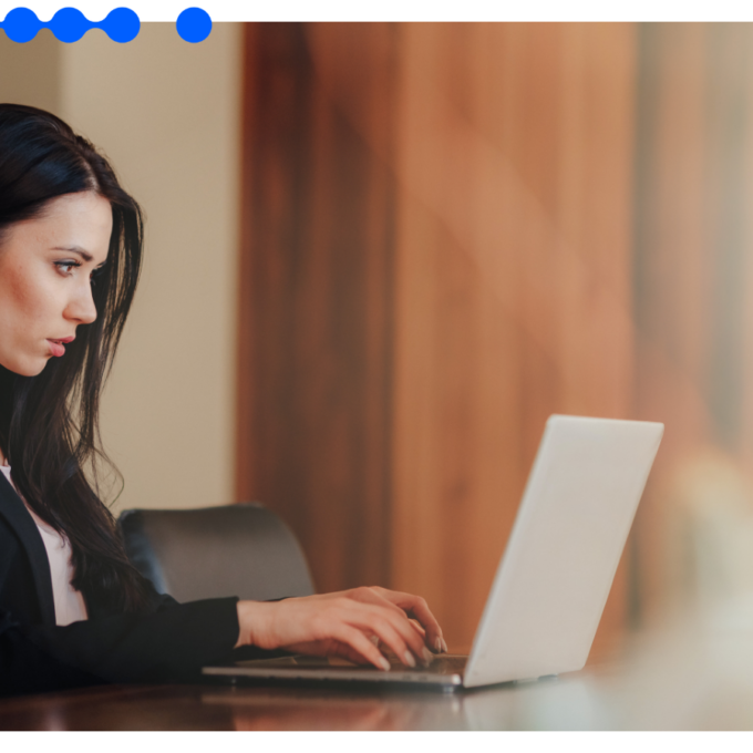 Young professional woman with long dark hair and a black blazer focused while working on a silver laptop at a wooden table, symbolizing the application of Embedded AI in business process orchestration