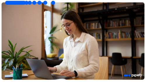 Mulher jovem de óculos e roupa branca sorrindo enquanto trabalha em um laptop em uma mesa de madeira com livros ao fundo, otimizando o fluxo de RH