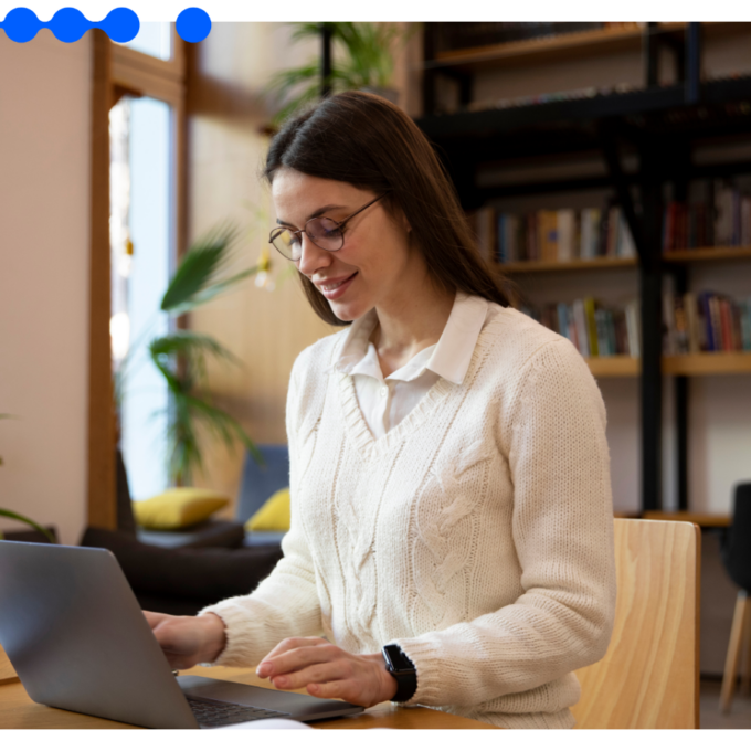 Mulher jovem de óculos e roupa branca sorrindo enquanto trabalha em um laptop em uma mesa de madeira com livros ao fundo, otimizando o fluxo de RH