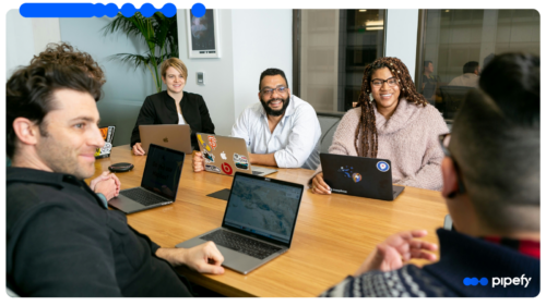 Grupo de profissionais sorrindo e conversando de forma descontraída ao redor de uma mesa de madeira com laptops em um escritório moderno, participando de uma reunião de onboarding de colaboradores