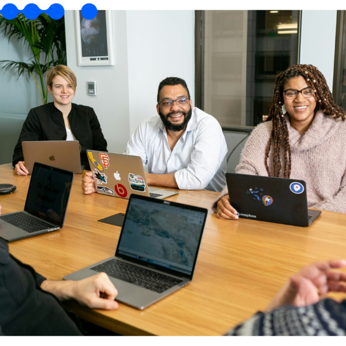 Grupo de profissionais sorrindo e conversando de forma descontraída ao redor de uma mesa de madeira com laptops em um escritório moderno, participando de uma reunião de onboarding de colaboradores
