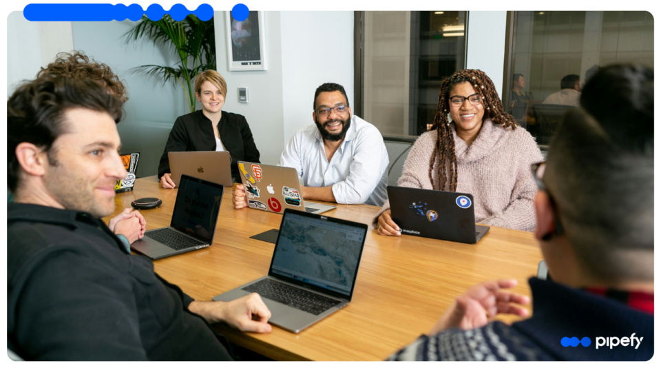 Grupo de profissionais sorrindo e conversando de forma descontraída ao redor de uma mesa de madeira com laptops em um escritório moderno, participando de uma reunião de onboarding de colaboradores