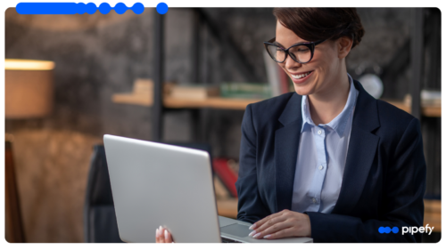 Smiling professional woman wearing glasses and a dark blazer, working on a laptop in a modern office, researching how to standardize processes