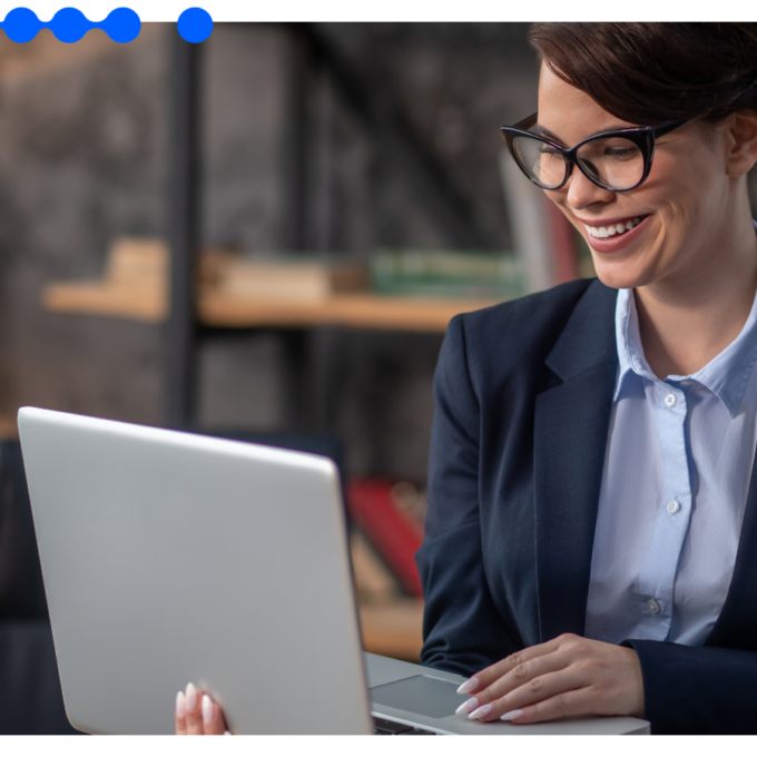 Smiling professional woman wearing glasses and a dark blazer, working on a laptop in a modern office, researching how to standardize processes