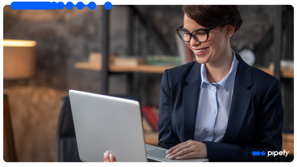 Smiling professional woman wearing glasses and a dark blazer, working on a laptop in a modern office, researching how to standardize processes