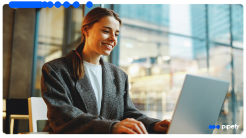 Mulher jovem de blazer cinza sorrindo enquanto trabalha em um laptop em um escritório moderno e iluminado, realizando a gestão de documentos