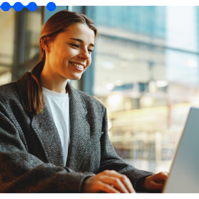 Mulher jovem de blazer cinza sorrindo enquanto trabalha em um laptop em um escritório moderno e iluminado, realizando a gestão de documentos