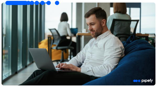 Homem trabalha em laptop sentado em puff azul em escritório corporativo moderno, com colega ao fundo, operando uma plataforma de orquestração com IA