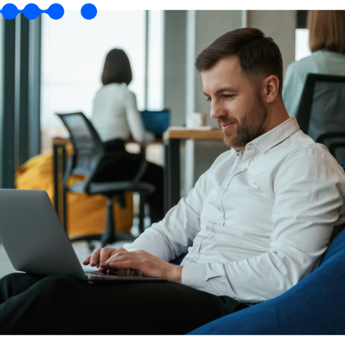 Homem trabalha em laptop sentado em puff azul em escritório corporativo moderno, com colega ao fundo, operando uma plataforma de orquestração com IA
