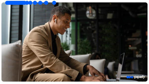 Young professional in a beige blazer smiling while typing on a laptop in a modern office lounge, with the Pipefy logo in the corner
